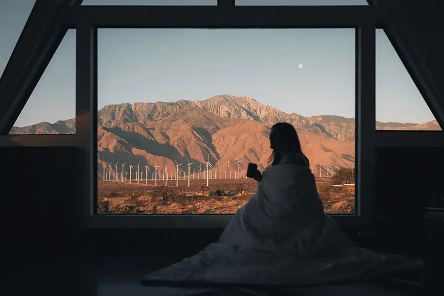 Silhouette of a woman, holding a mug, looks out an oversized window at brown, desert mountains