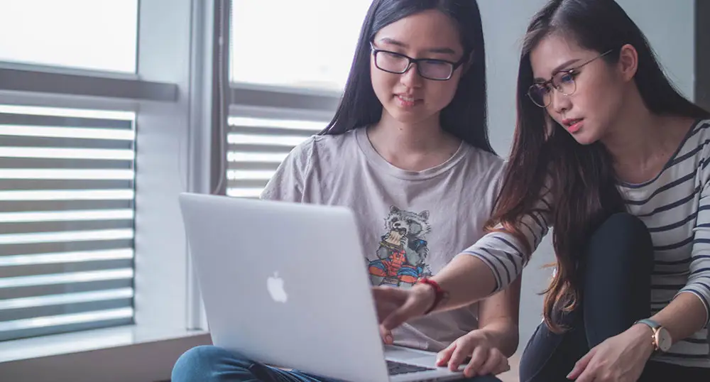 Two young women use a laptop together by a window