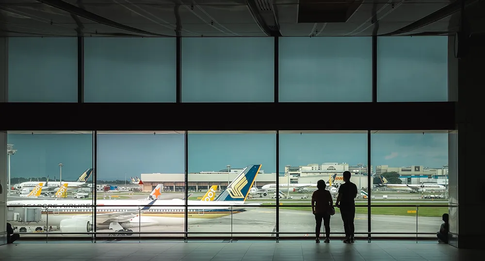 Two people stand at window in airport, looking out at airplanes