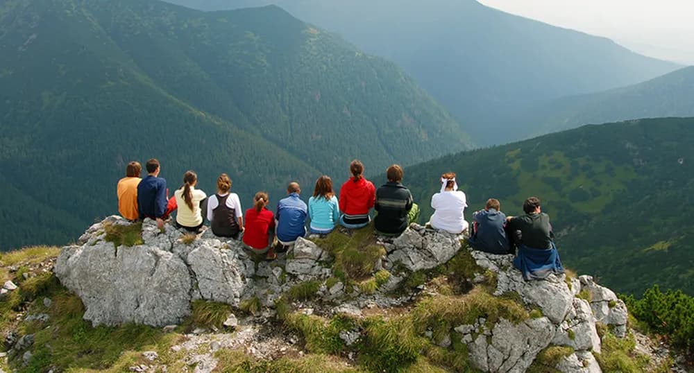 Large group of people sit on a rock outcropping, looking at mountains