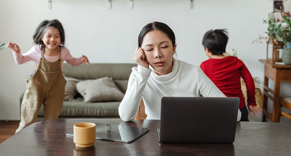 Exhausted-looking woman stares at laptop while kids run around in background